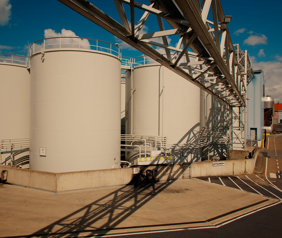 Storage tanks in Antwerp, caught under a hard blue July sky with a pipe gantry throwing long diagonals across the concrete.